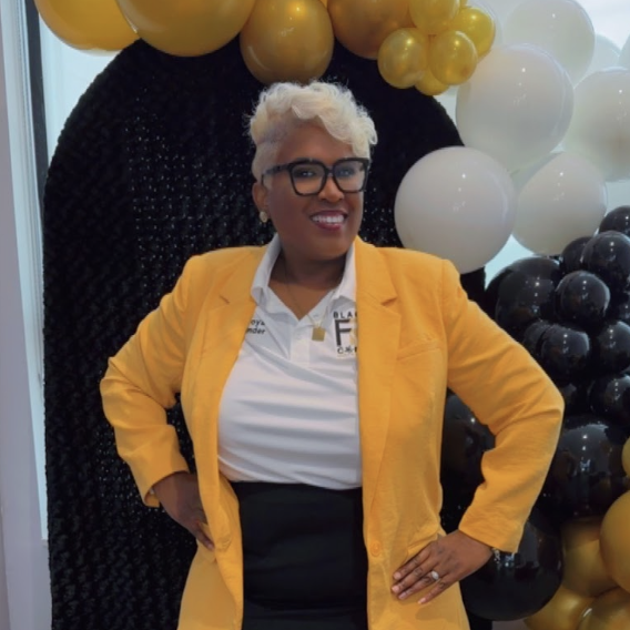 Founder Latoya Wise, known as Toya Imani, in a mustard blazer and Black FRMs Connect polo standing confidently in front of the community's black, gold and white balloon arch